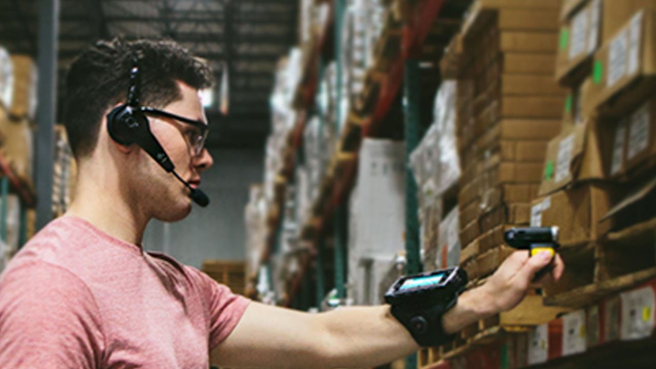 A male warehouse worker uses a headset, wearable mobile computer on his wrist and a handheld scanner to confirm inventory on a shelf.
